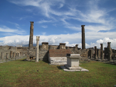 Photo shows the column with the sundial in front of the remains of a stone wall, with rows of columns on each side of the wall.